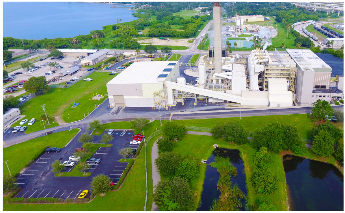 Aerial view of an industrial facility near a lake, surrounded by greenery and parked cars.