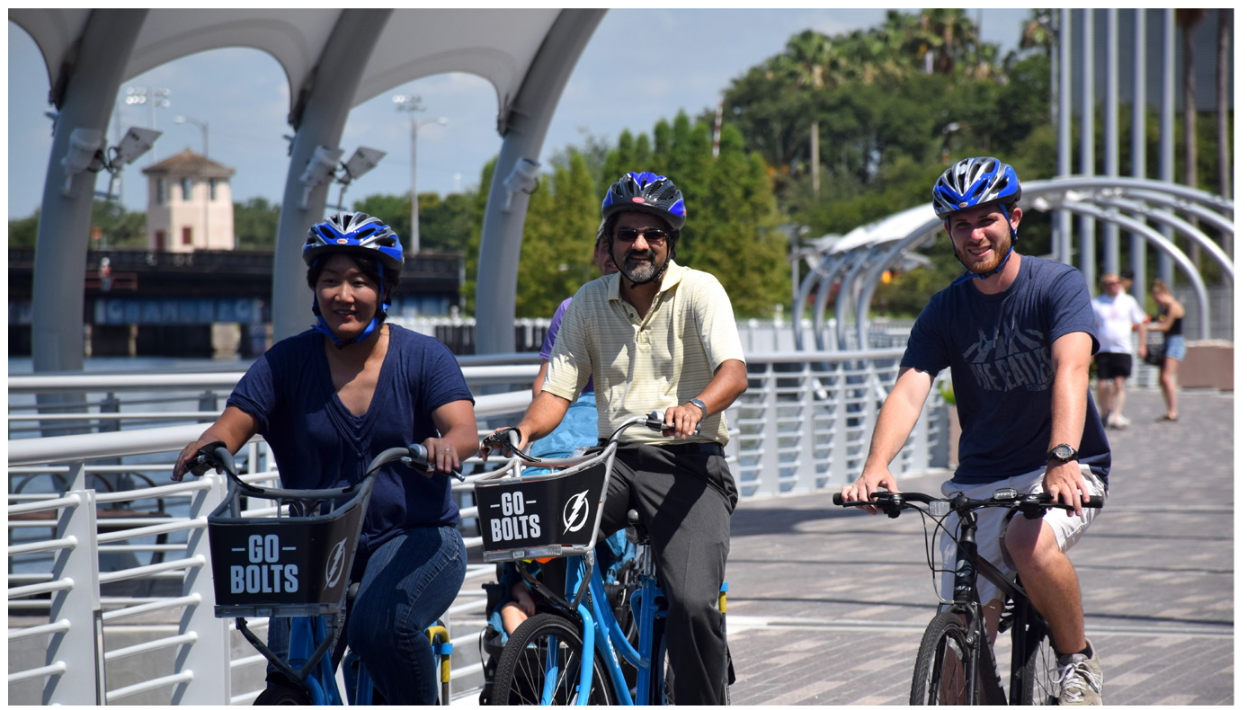 Three people riding bicycles on a walkway, wearing helmets, with blue bikes labeled 'Go Bolts'.