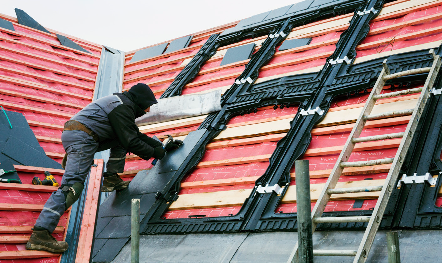 A roofer installing black shingles on a sloped roof, with wooden support and ladders visible.