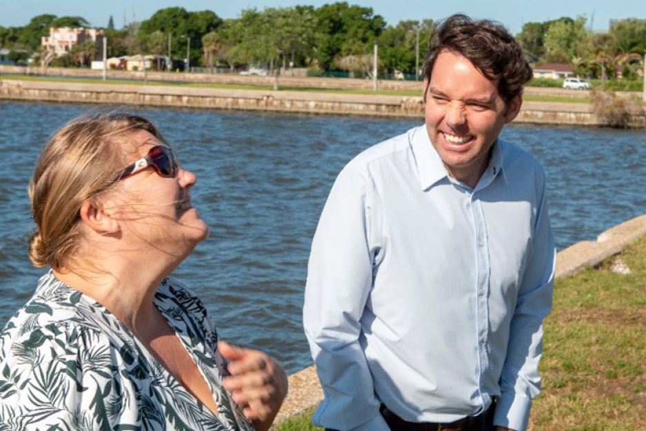 Two people laughing and talking by a waterfront, with greenery and buildings in the background.