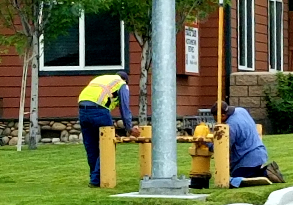 Two workers in vests inspecting a yellow fire hydrant on a grassy area next to a building.