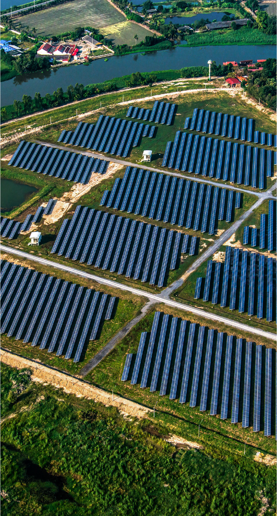 Aerial view of a large solar panel farm set in a green landscape with water bodies in the background.