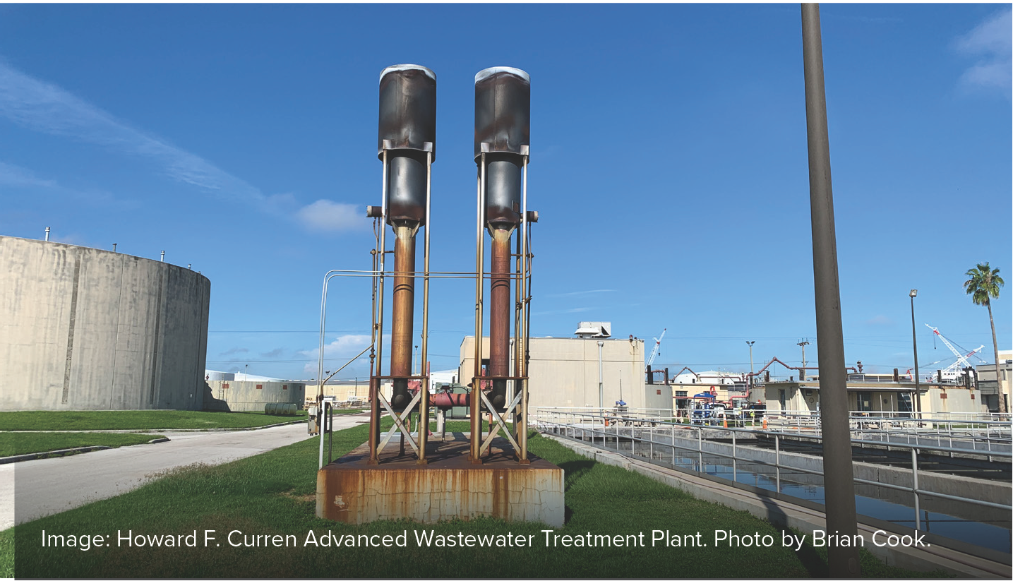 Two tall water towers next to a wastewater treatment plant with storage tanks and a clear blue sky.