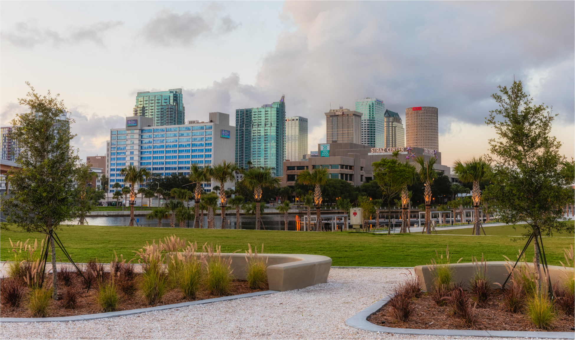 View of a city skyline with modern buildings and palm trees in a park setting during sunset.