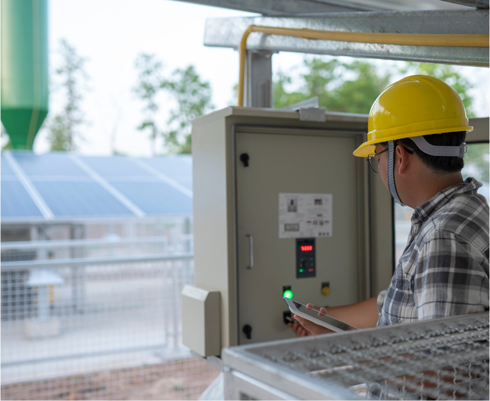 A worker in a yellow hard hat operates a control panel at a facility with solar panels in the background.