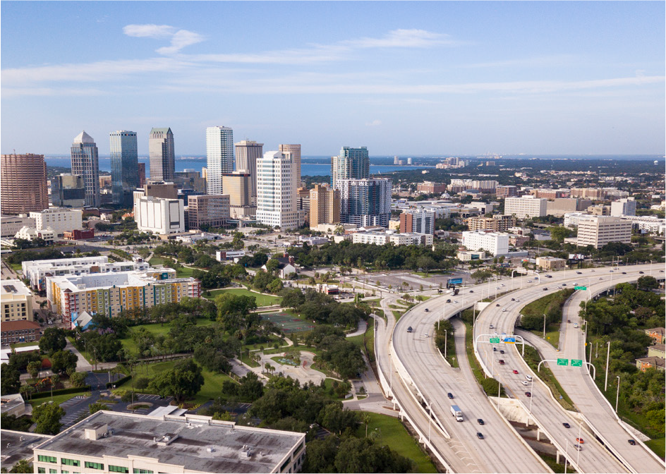 Aerial view of a city skyline with tall buildings, green spaces, and highways under a clear blue sky.