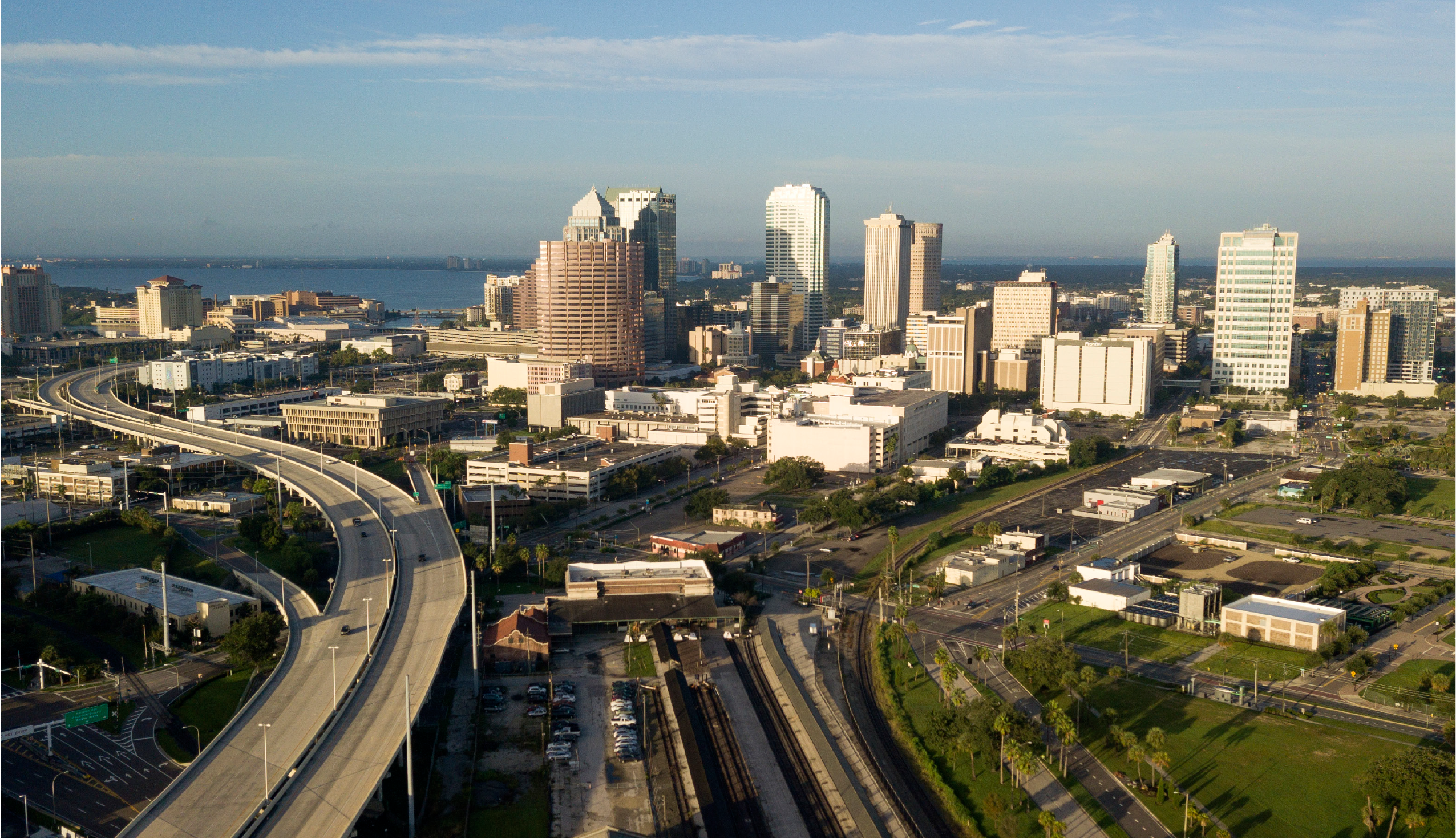 Aerial view of a city skyline with skyscrapers, highways, and waterfront, under a clear blue sky.