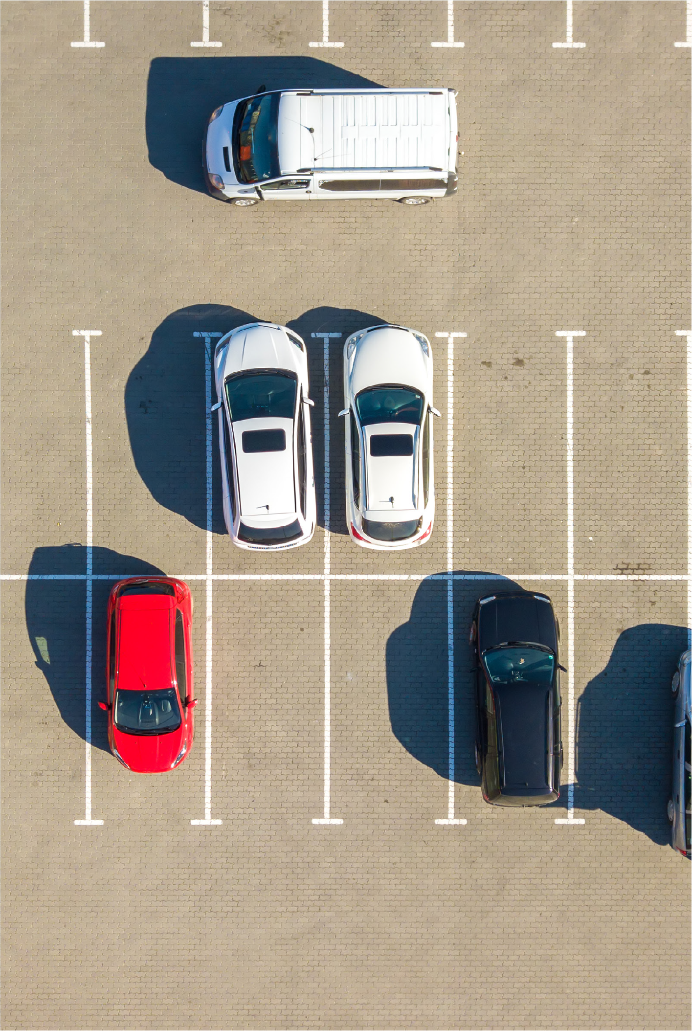 Aerial view of a parking lot with seven parked cars, including a red sedan, two white sedans, and a black sedan, among empty spaces.