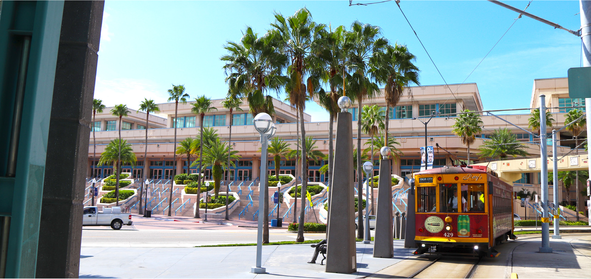 A vintage tram on a street lined with palm trees, in front of a modern building and stairs.