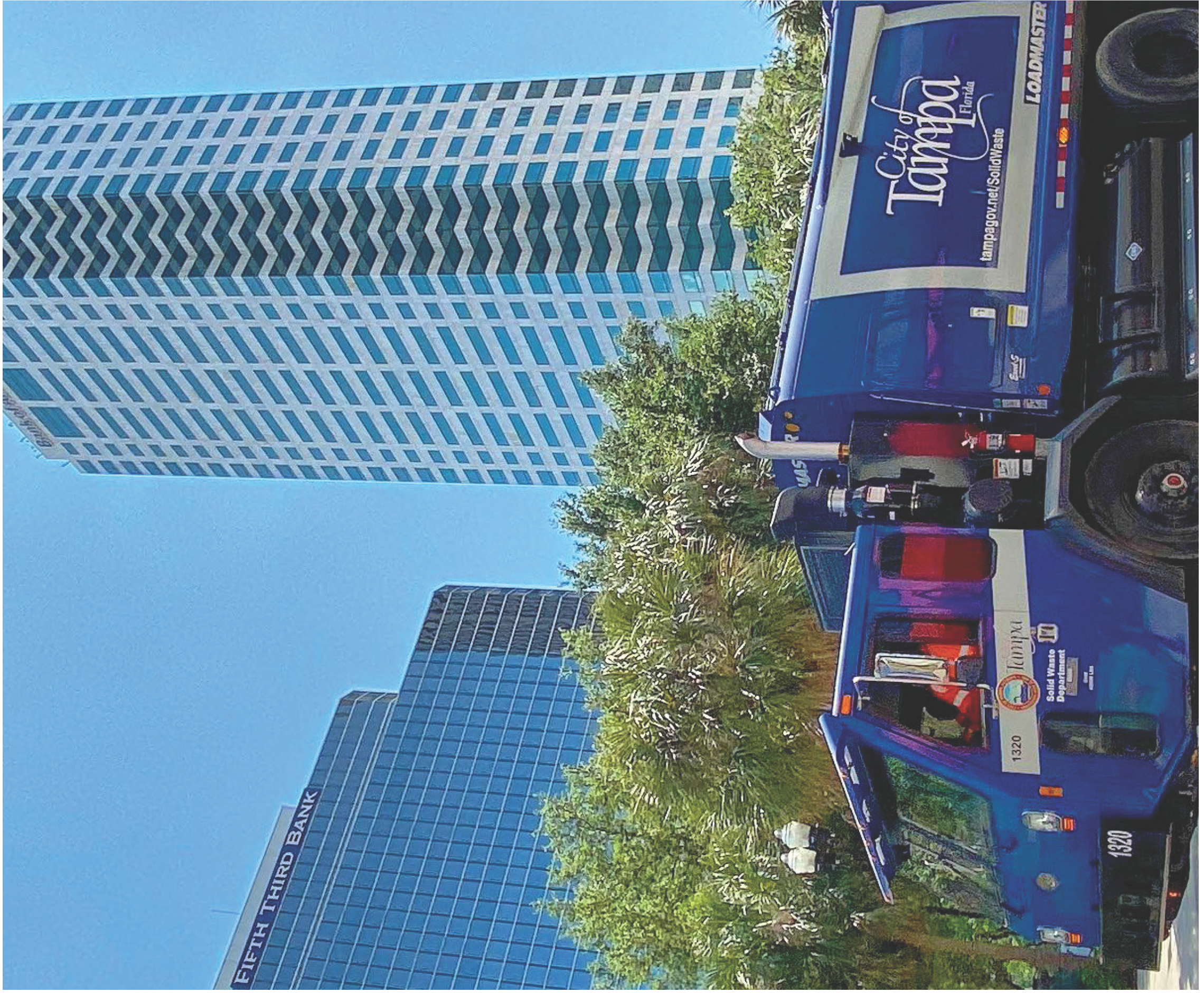 Two service trucks parked in front of tall buildings under a clear blue sky, surrounded by greenery.