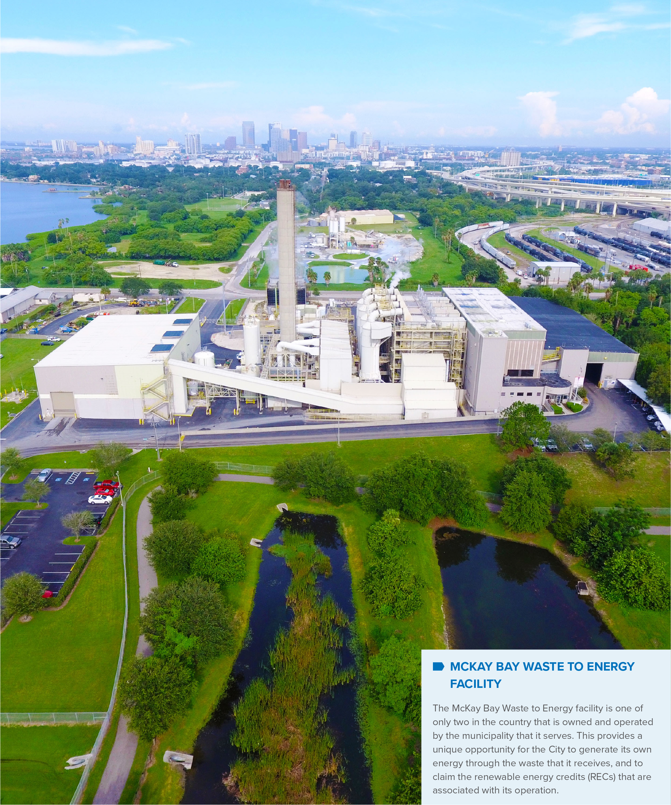 Aerial view of the McKay Bay Waste to Energy Facility, surrounded by greenery and water, with a city skyline in the background.