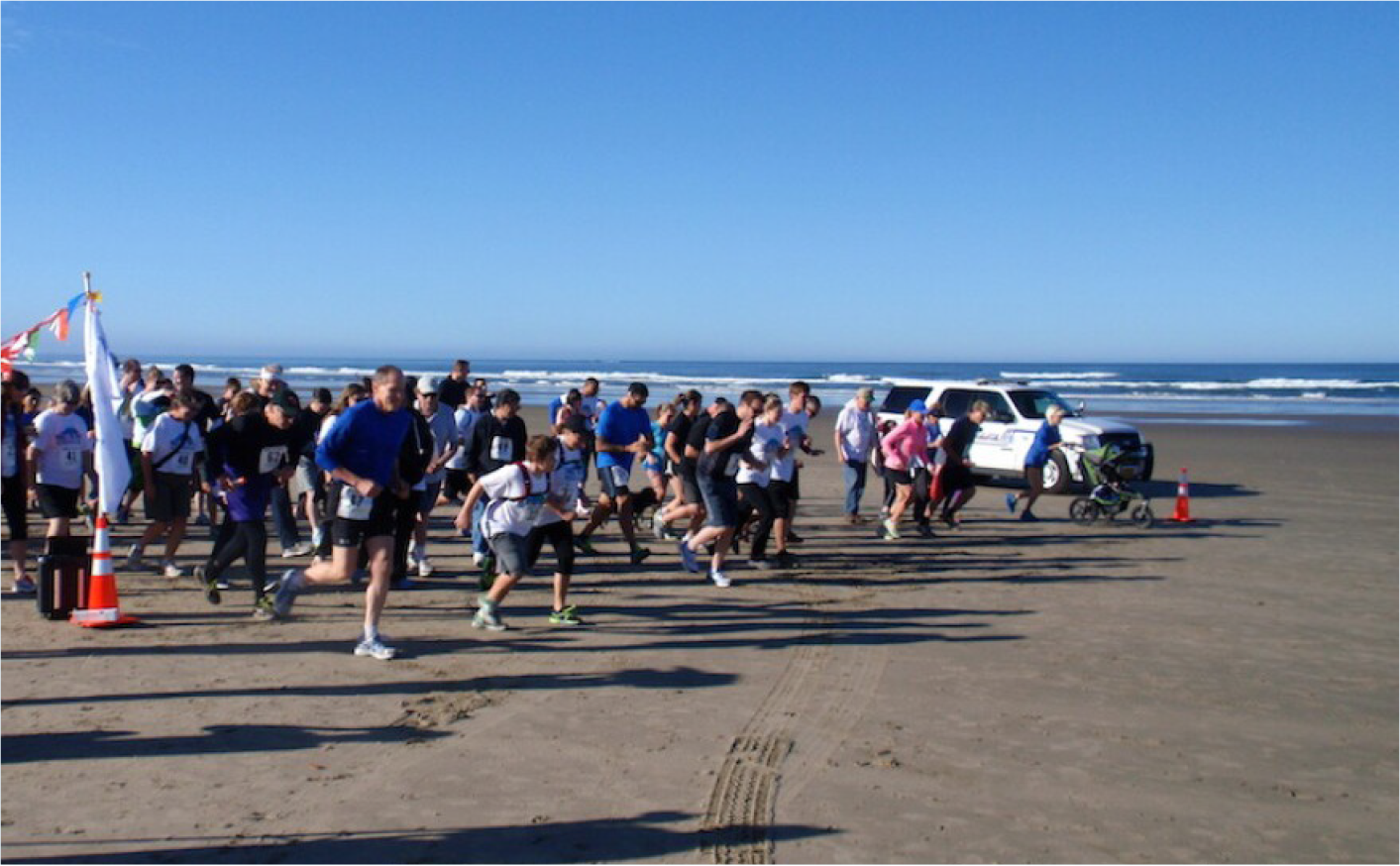 A group of runners starting a race on a sandy beach with ocean waves in the background and a police vehicle nearby.