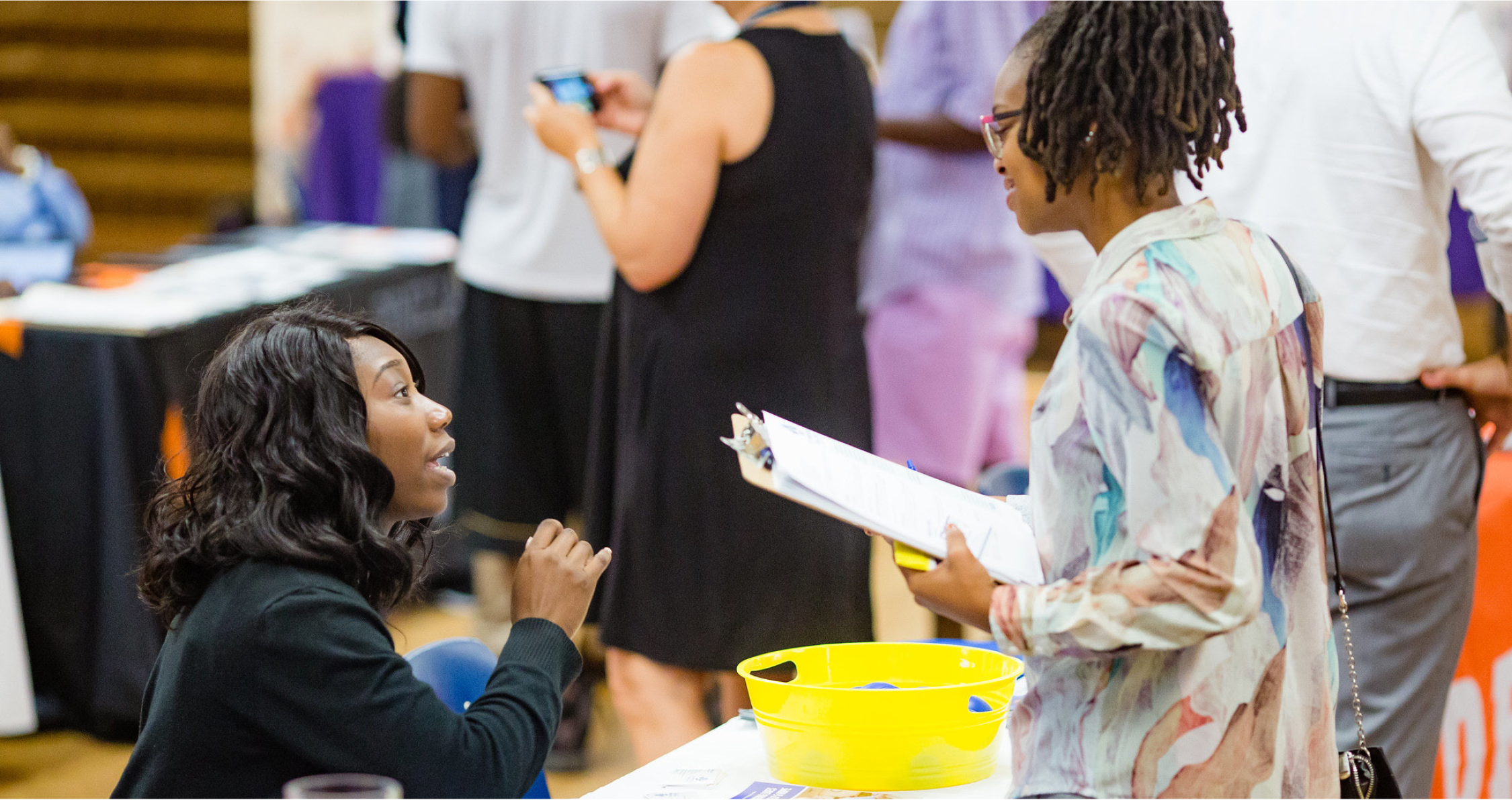 A woman talks to another woman holding a clipboard at an event, with people in the background.