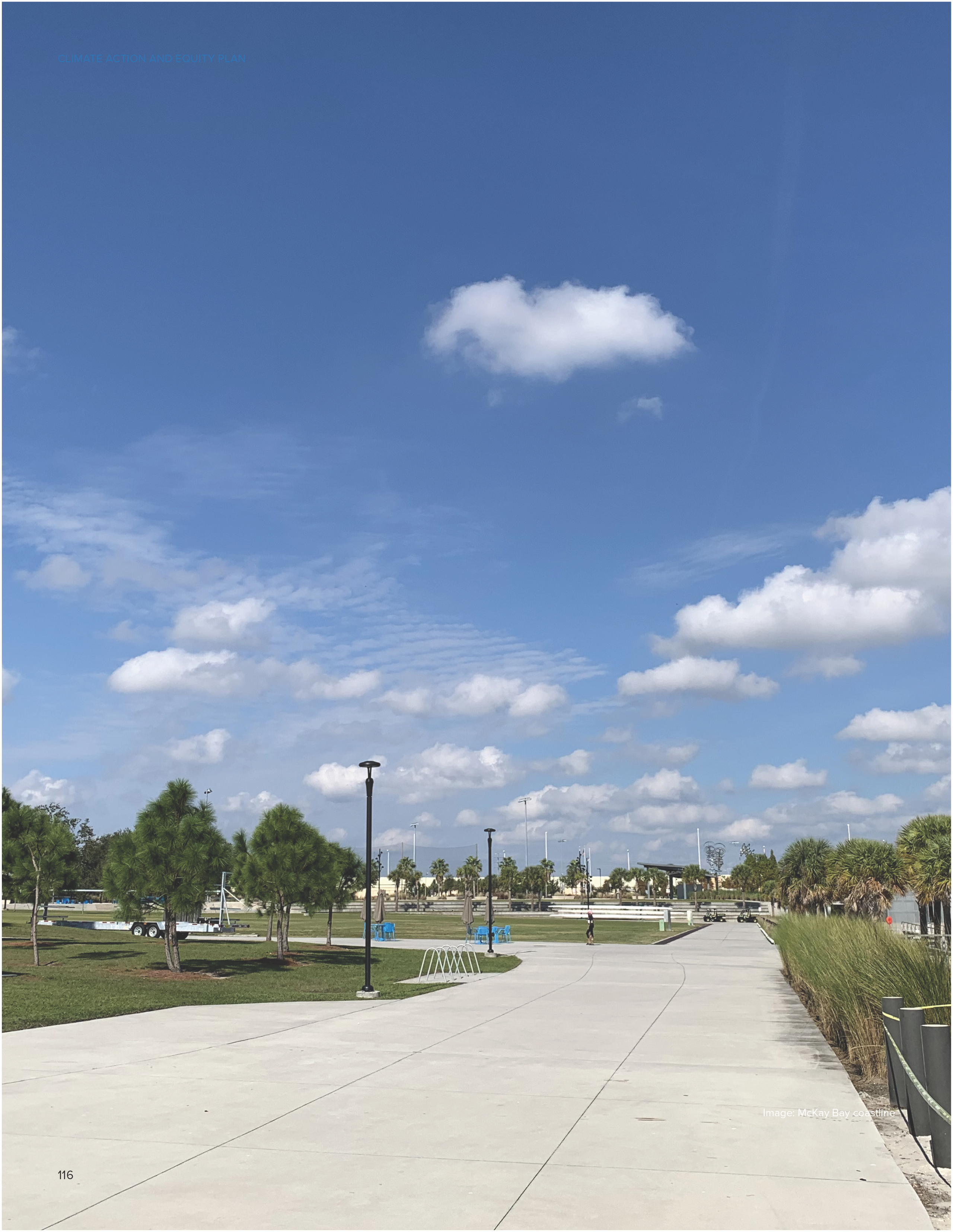 A wide walkway lined with small trees, under a blue sky with fluffy clouds, leading towards buildings in the distance.