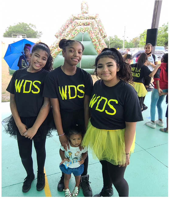 Three girls in black shirts with 'WDS' printed in yellow, wearing yellow tutus, with a toddler in front at an outdoor event.