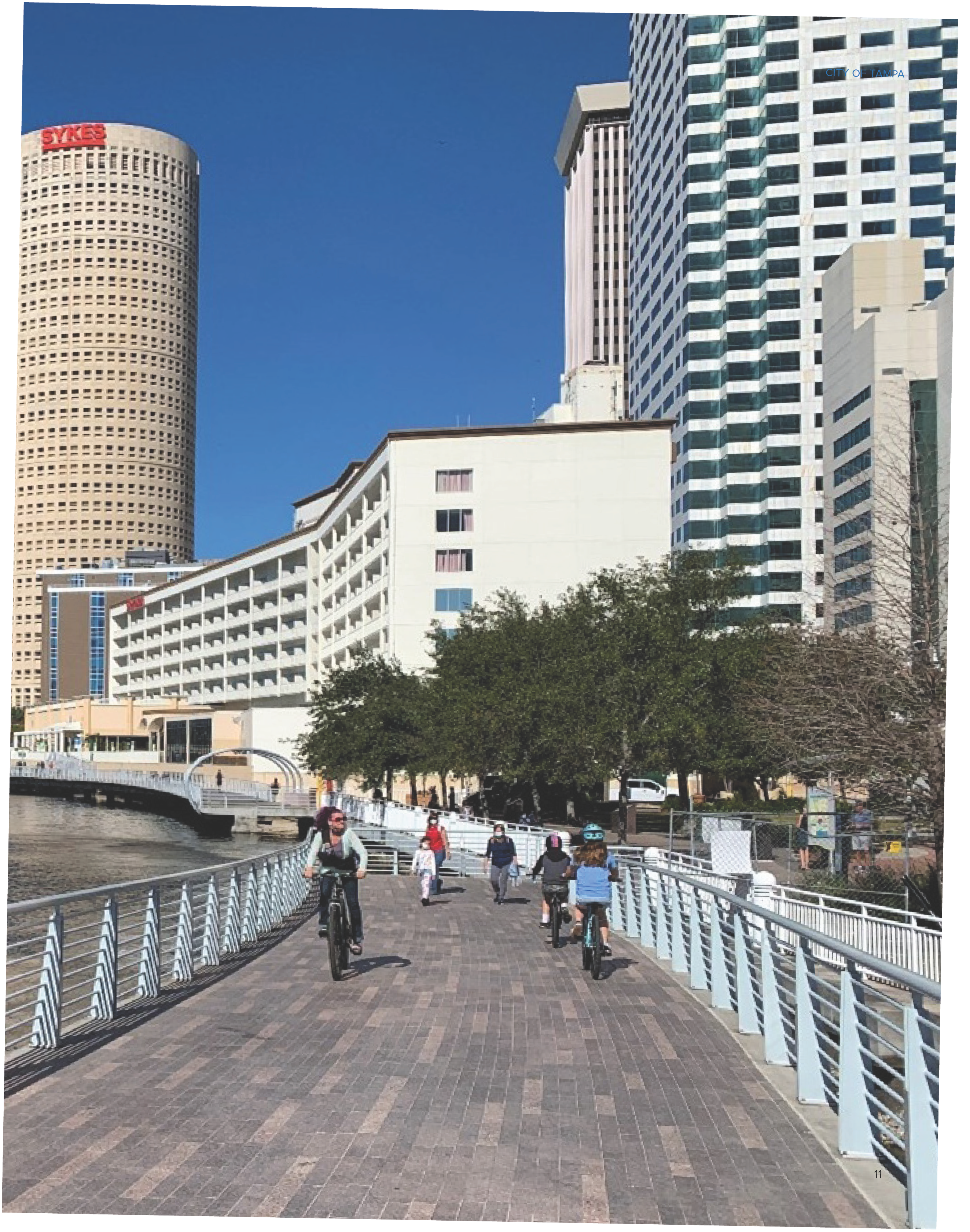 People cycling and walking along a riverside path with tall buildings in the background under a clear blue sky.