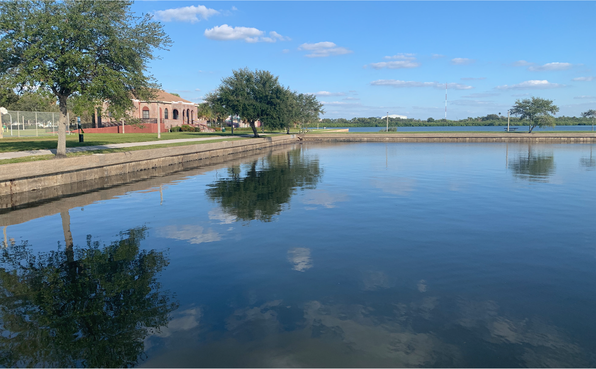 Serene water reflection of trees and a building along a peaceful lakeside setting under a clear blue sky.