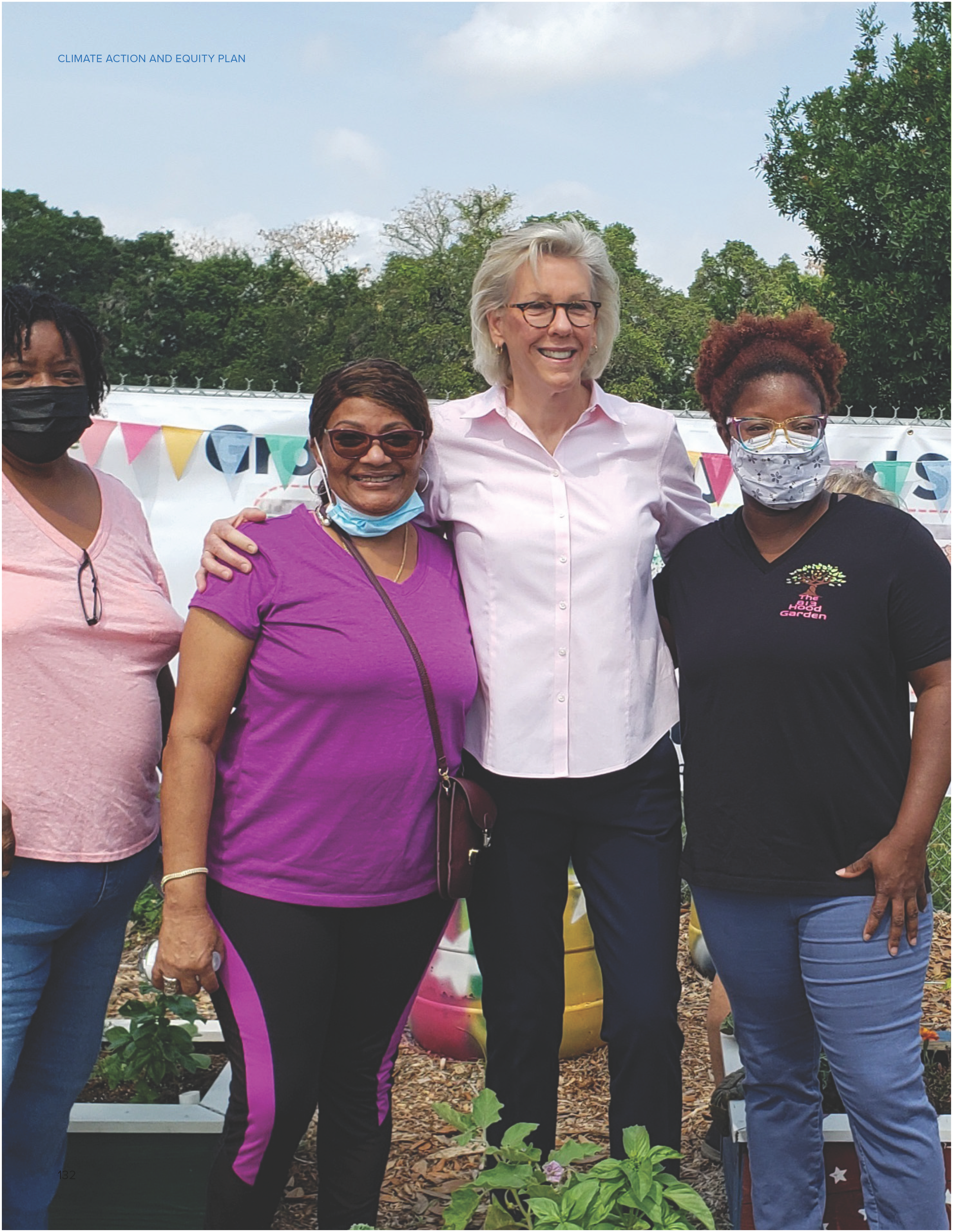 A group of four women, smiling, standing together in a garden setting, wearing masks and casual attire.