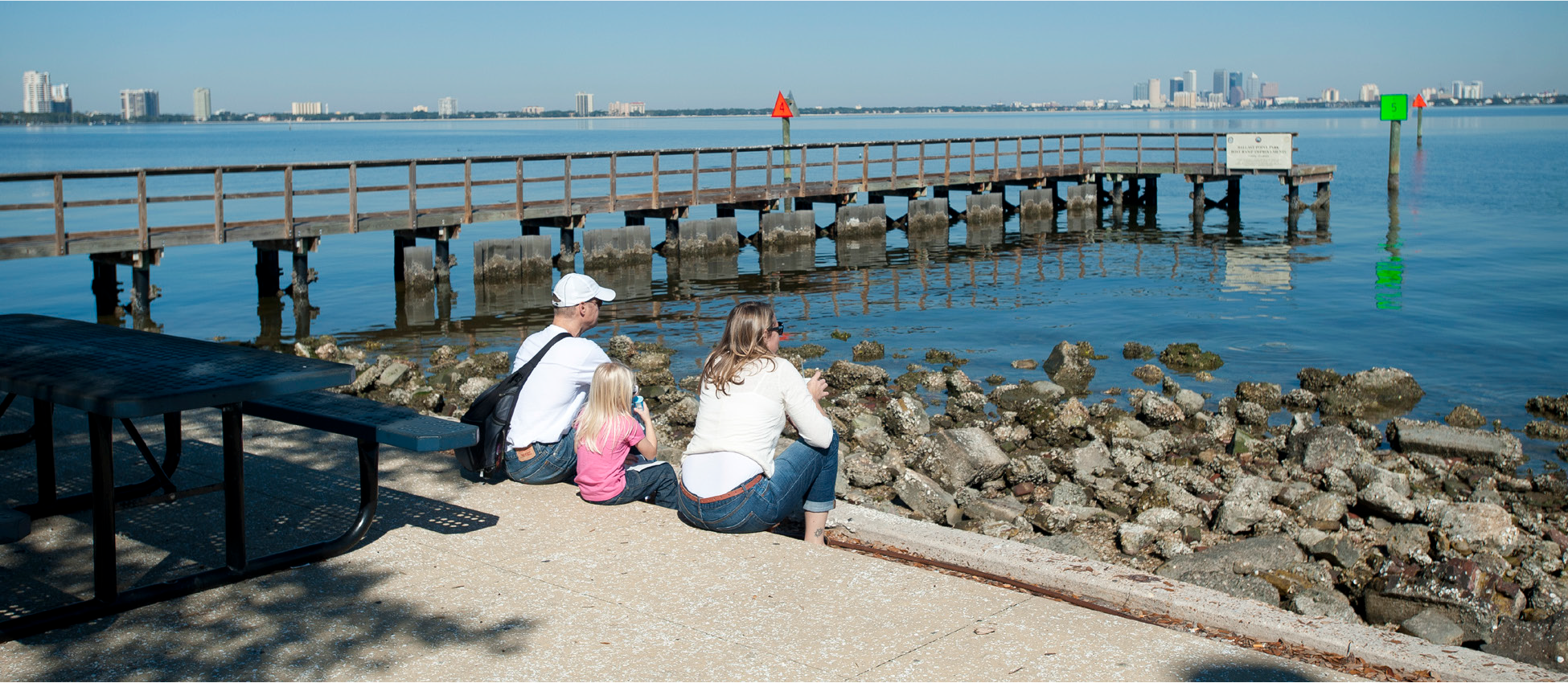 A family sits on a shore near a calm water body, observing a pier and city skyline in the background.
