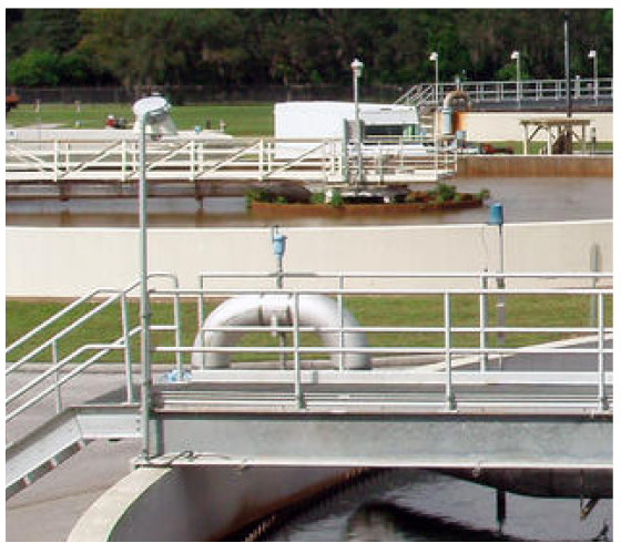 An industrial water treatment facility with metal walkways, pipes, and tanks, surrounded by green grass and trees.