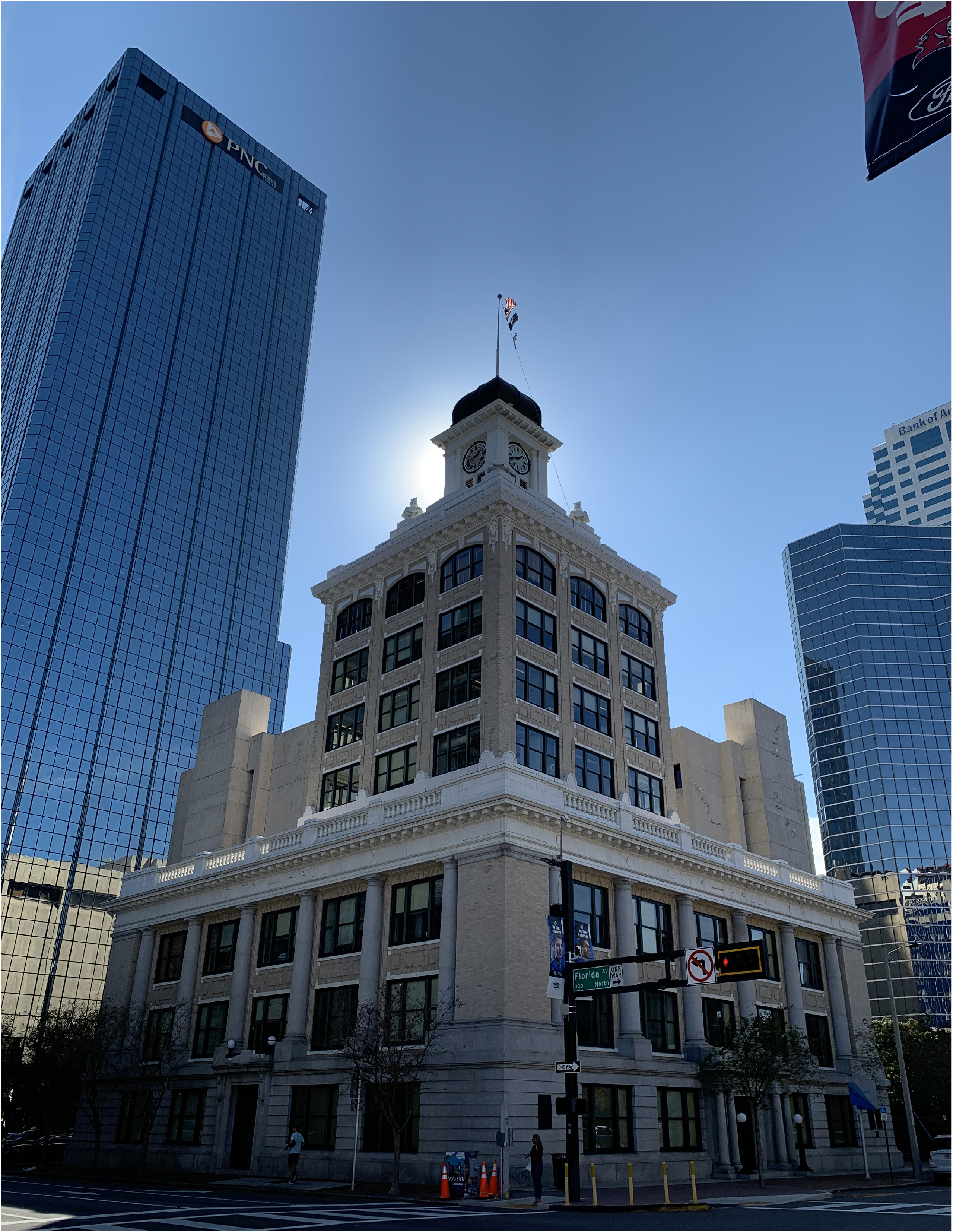 Historic building with a clock tower, surrounded by modern skyscrapers and bright sunlight in a cityscape.