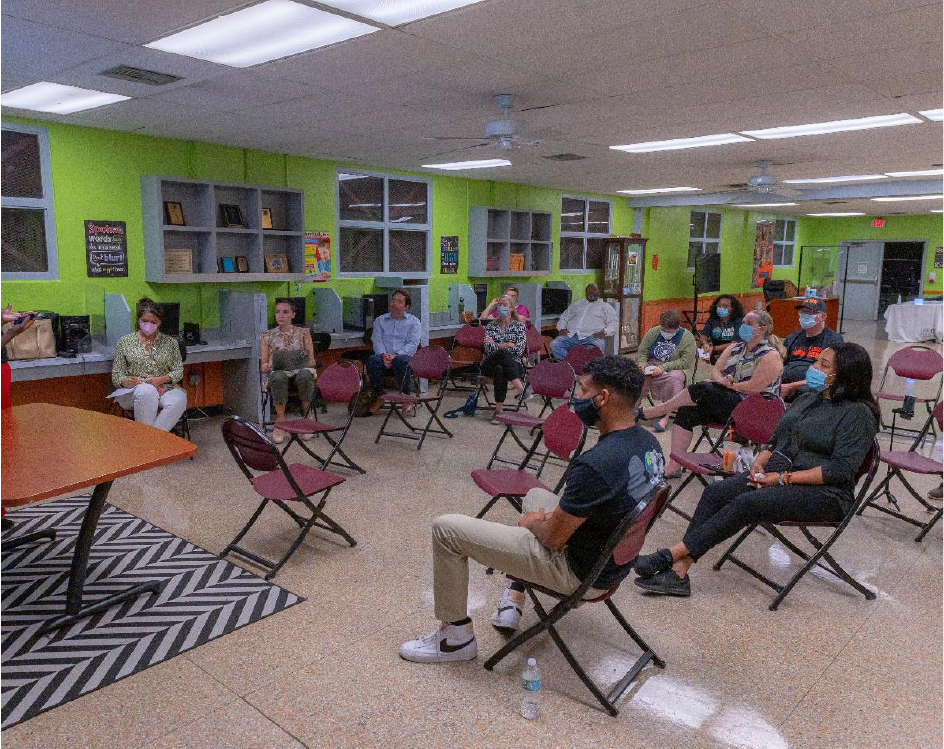 A group of people seated in a well-lit community room, wearing masks and maintaining distance, with green walls and tables in the background.