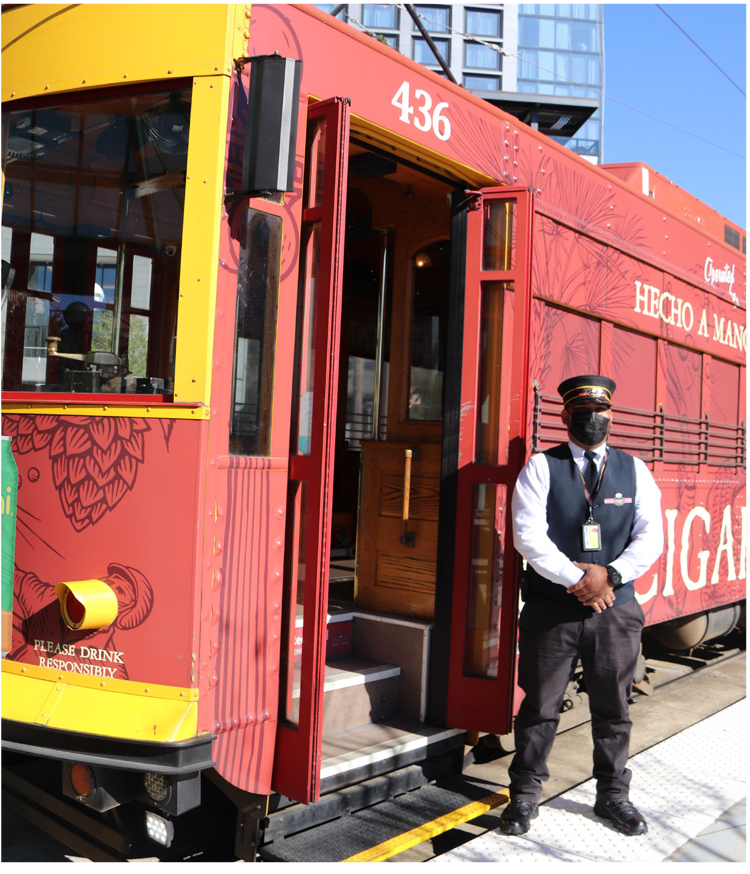 A streetcar with a conductor standing beside the open door, wearing a uniform and mask, in a vibrant urban setting.