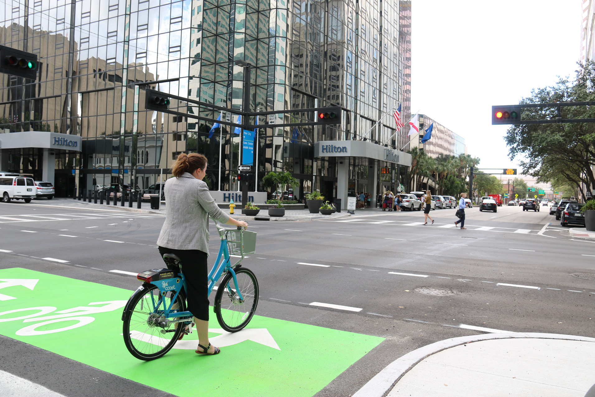 A person on a bike waits at an intersection with traffic signals, in front of a Hilton hotel and glass buildings.