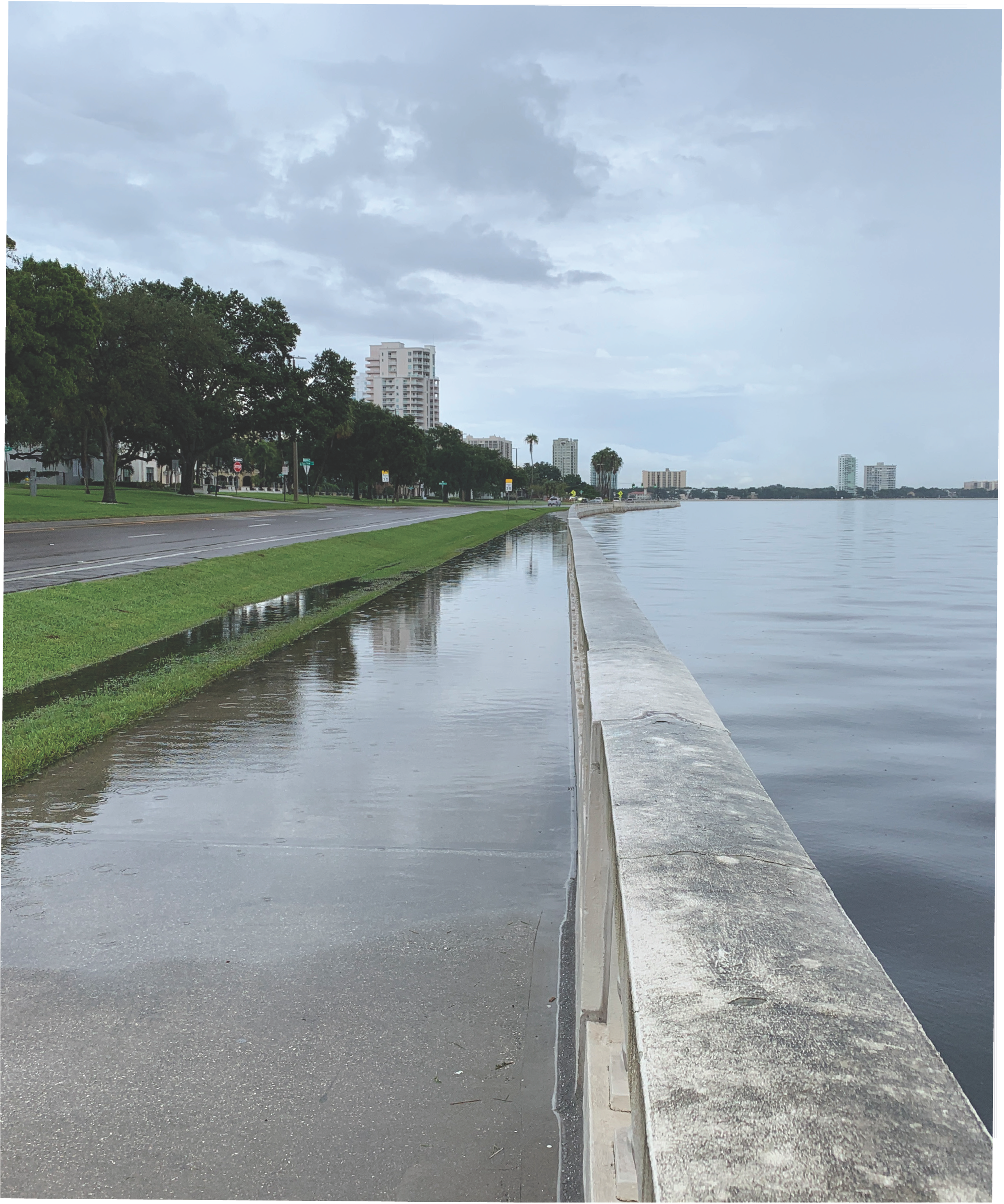A waterfront view with a calm lake, overcast sky, and grass-lined pathway; reflecting buildings and trees along the shore.