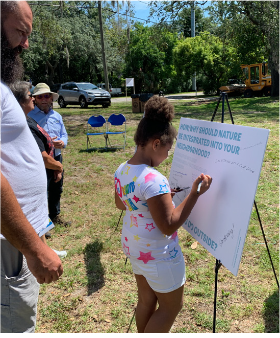 A young girl writes on a whiteboard during a community event about integrating nature into neighborhoods, with people observing nearby.