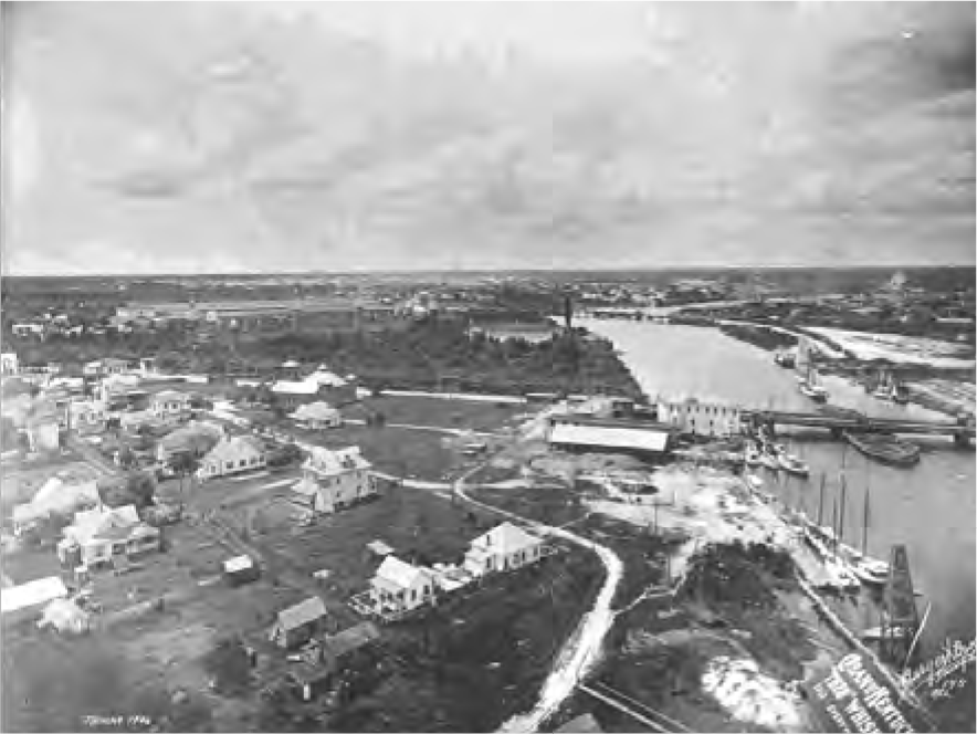 Aerial view of a riverside town in the early 1900s, featuring houses, a bustling river with boats, and a cloudy sky.