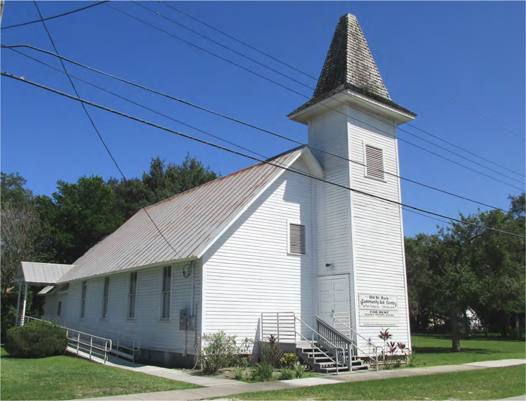 A white church building with a steeple, surrounded by green trees and power lines, under a clear blue sky.
