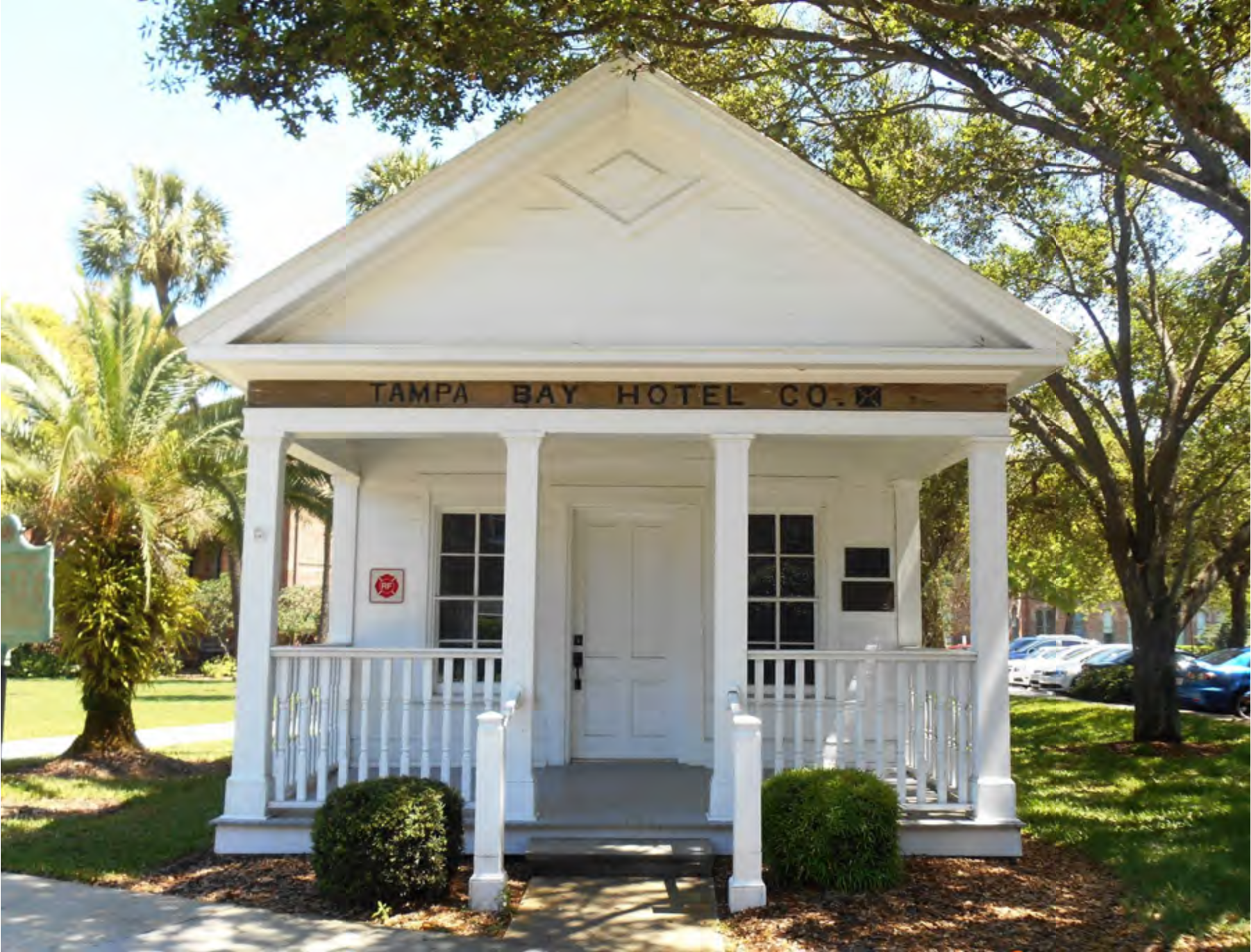 Small white building with wooden porch, labeled 'TAMPA BAY HOTEL CO.', surrounded by greenery and trees.