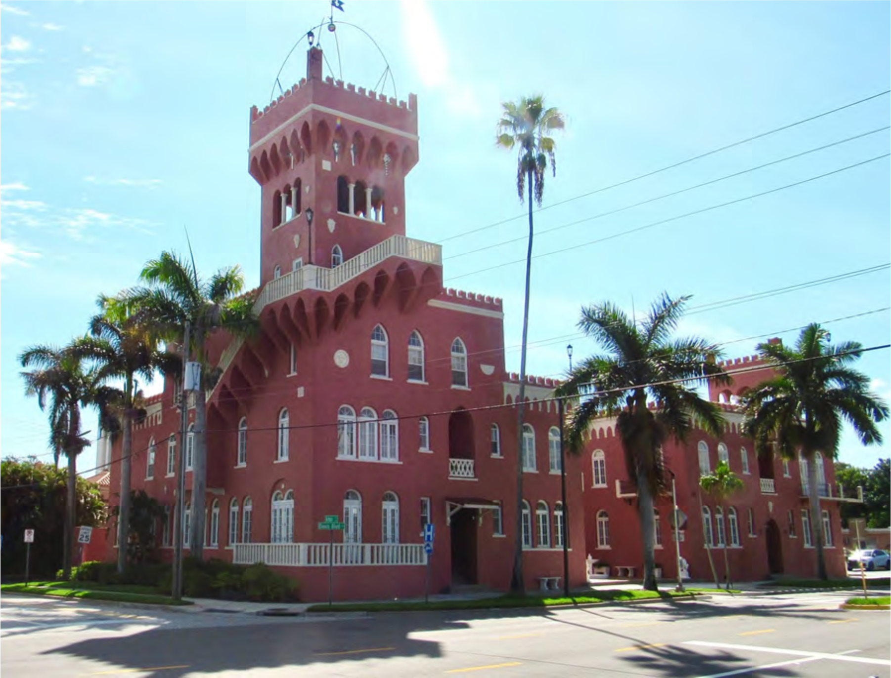 A red brick castle-like building with a tower, palm trees, and a clear blue sky in the background.