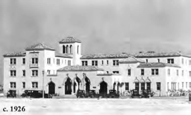 Historic building from 1926 featuring Spanish architecture, with arched windows and a bell tower, surrounded by old vehicles.
