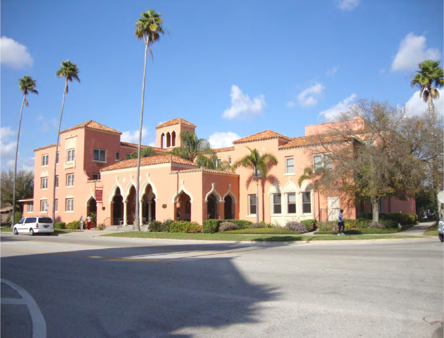 A pink Mediterranean-style building with palm trees, featuring arches and a prominent tower, situated at a street corner on a sunny day.