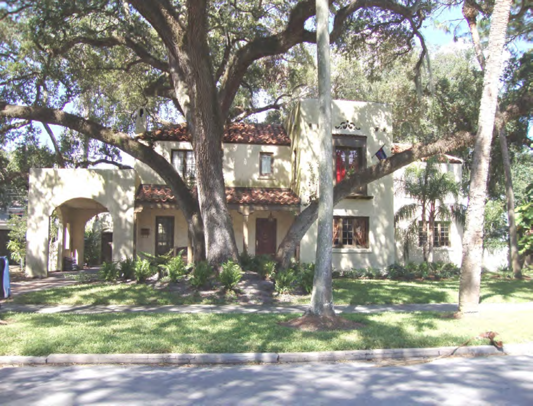 Two-story stucco house with a red tile roof, large oak tree in front, surrounded by landscaped greenery.
