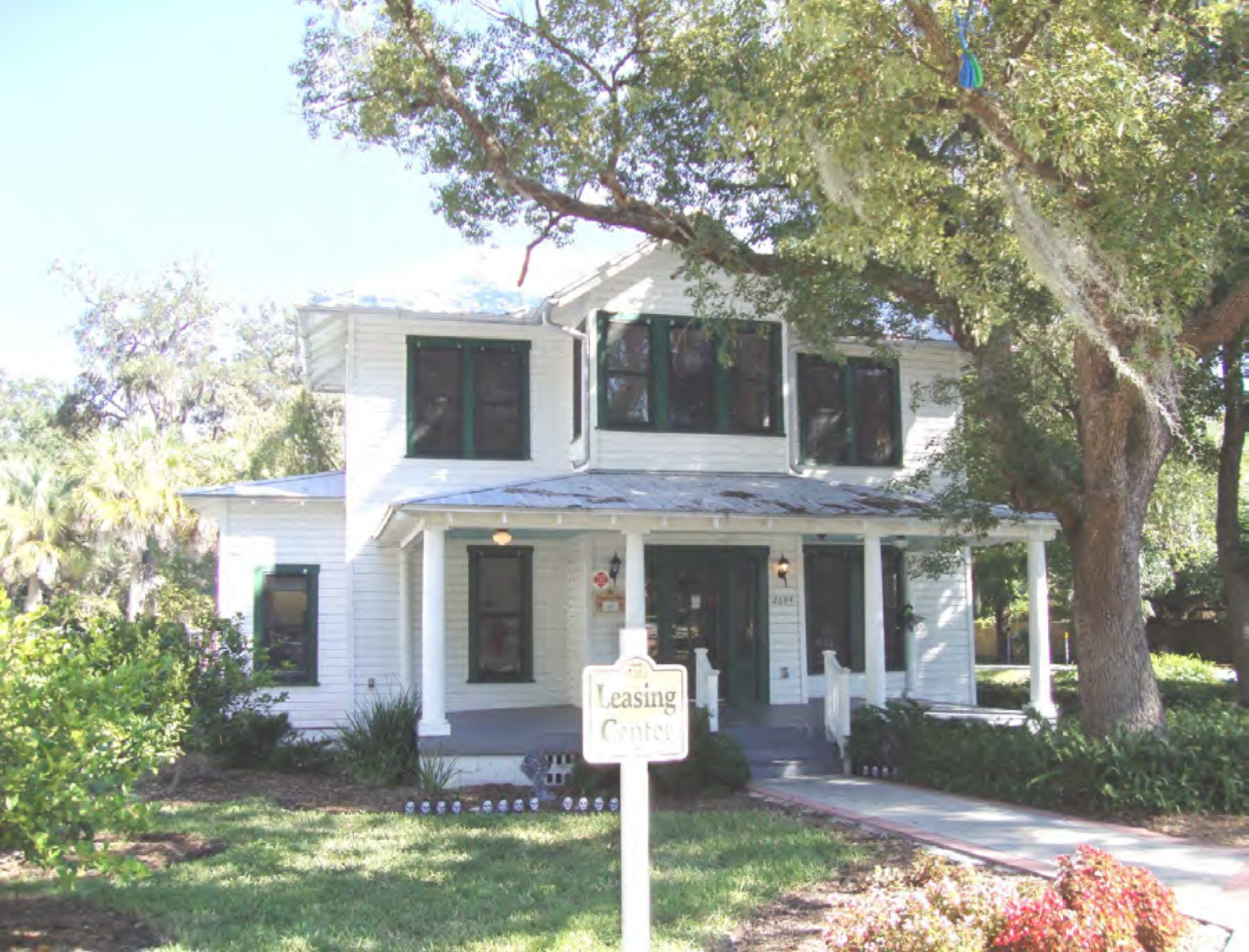A two-story white house with green shutters, surrounded by trees and landscaping, featuring a sign for leasing in the foreground.