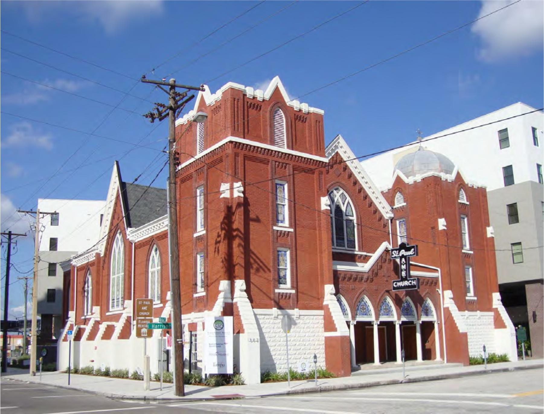 Brick church building with gothic architecture, featuring arched windows and a prominent steeple, surrounded by modern buildings and power lines.