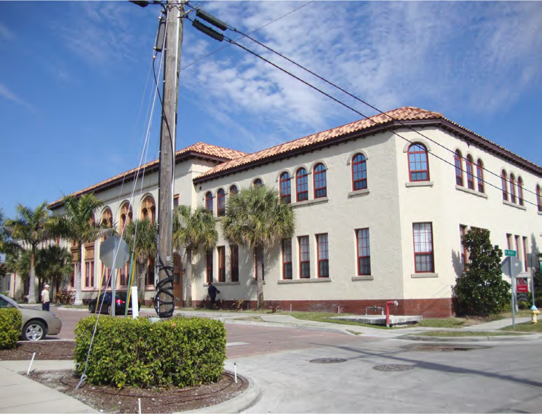 A two-story historical building with a tiled roof, surrounded by palm trees and power lines, located at a street intersection.