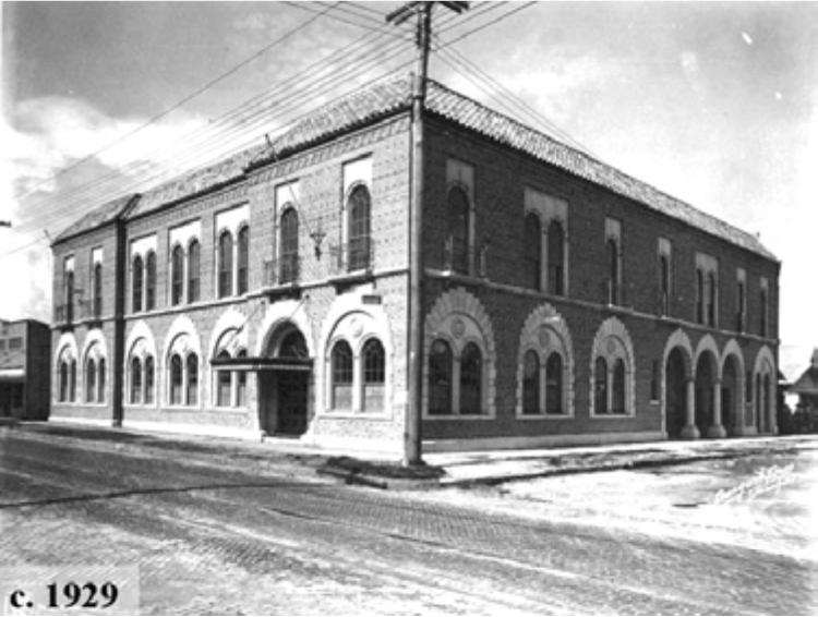 Historic building from approximately 1929, featuring arched windows and entrance, situated on a street corner.