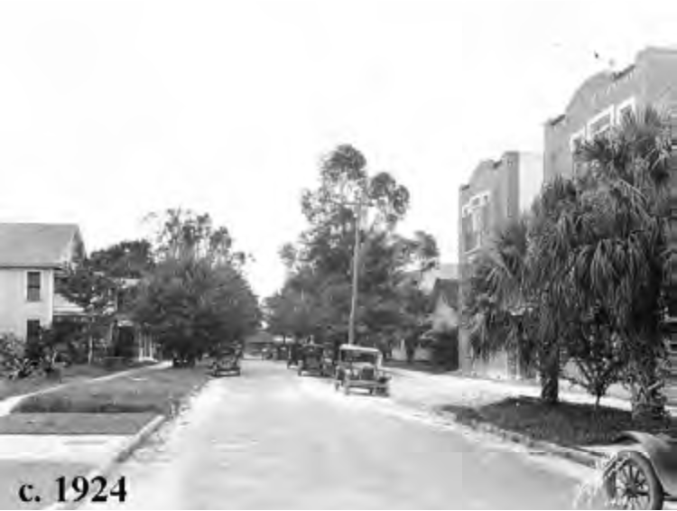 Historic street scene from c. 1924, featuring vintage cars and trees lining the road.
