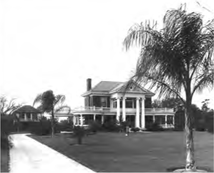 Large historic house with columns, surrounded by palm trees and a grassy lawn, along a pathway.