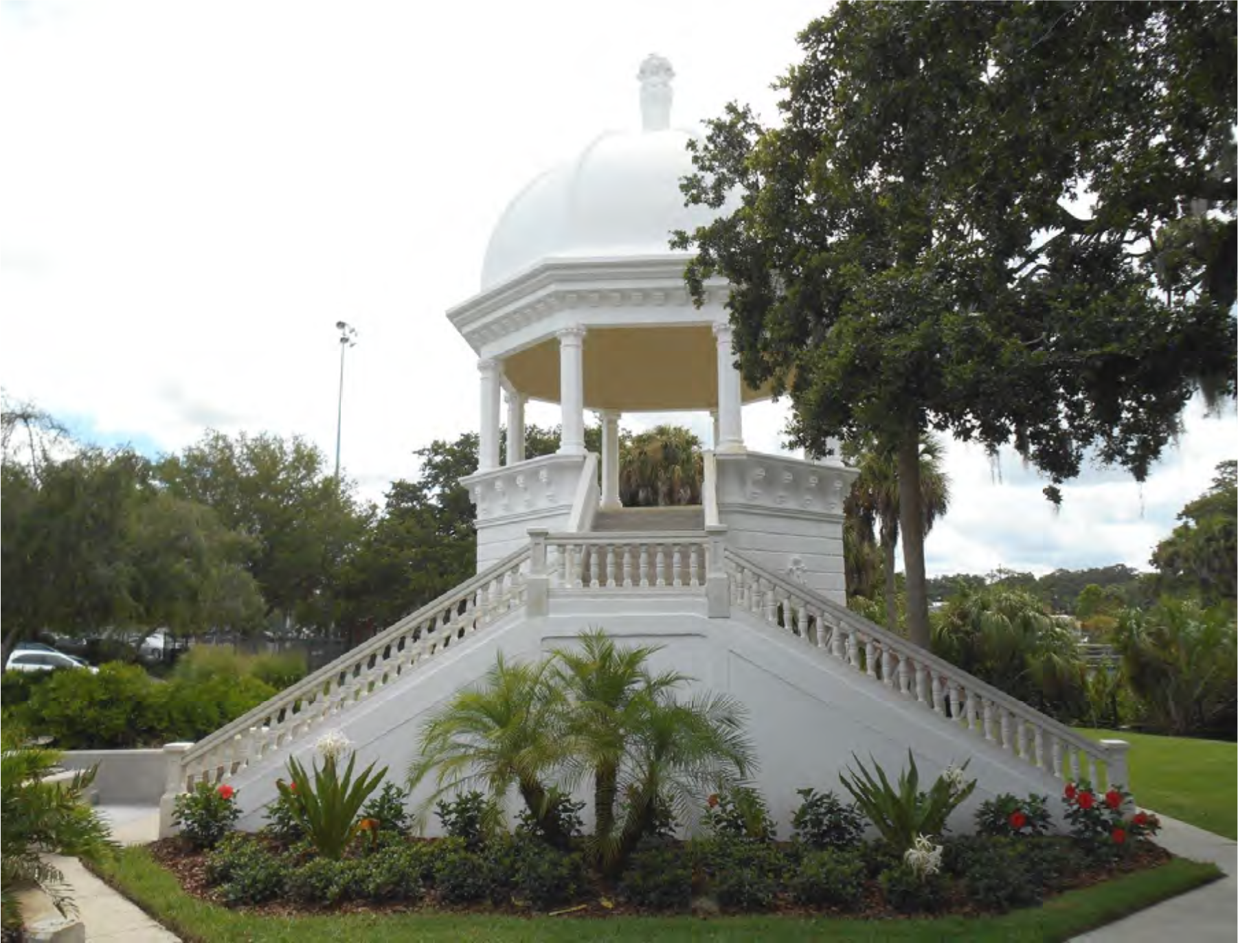 A white gazebo with a dome, surrounded by lush greenery and colorful flowers, set in a park.