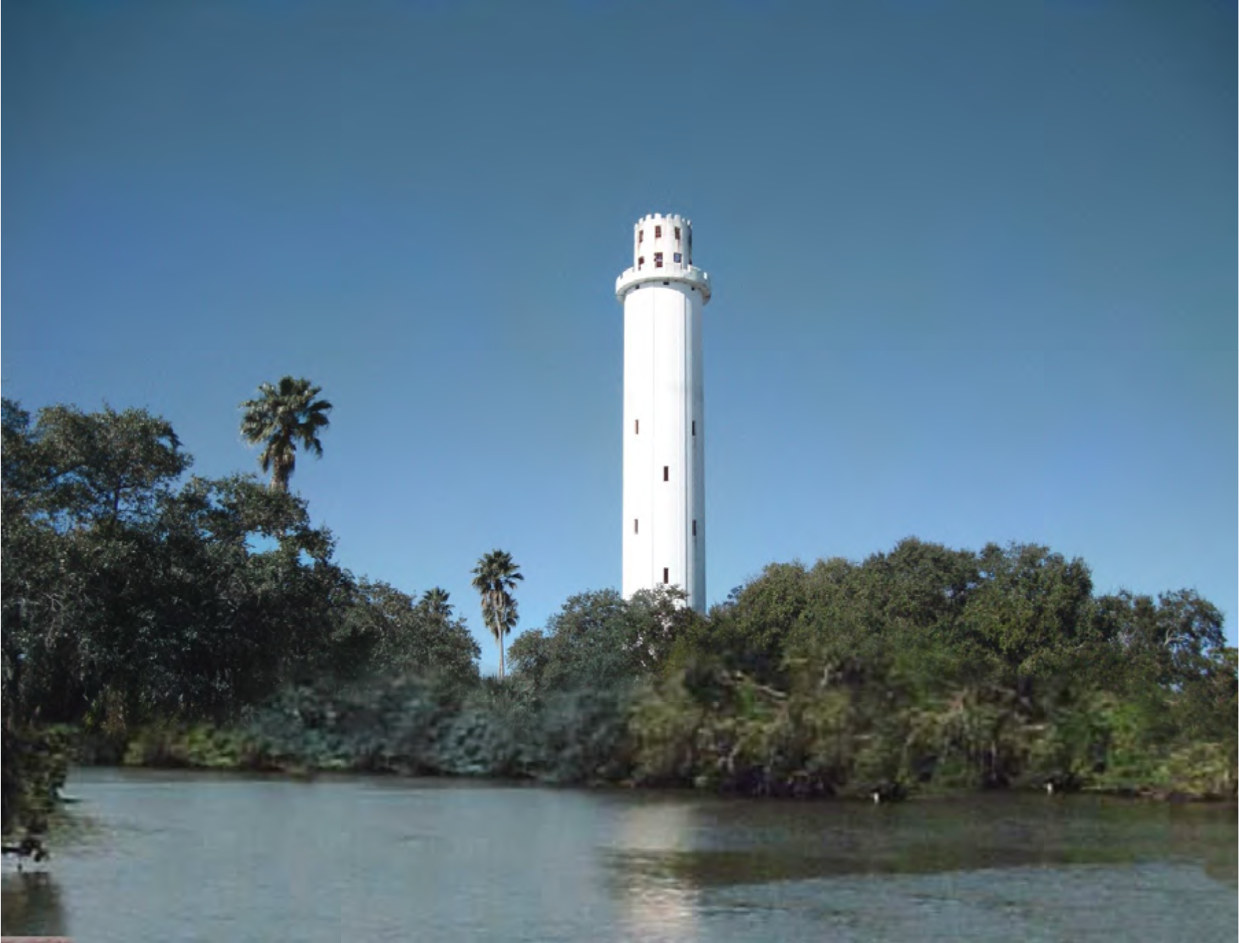 A tall white tower surrounded by lush greenery and a river under a clear blue sky.