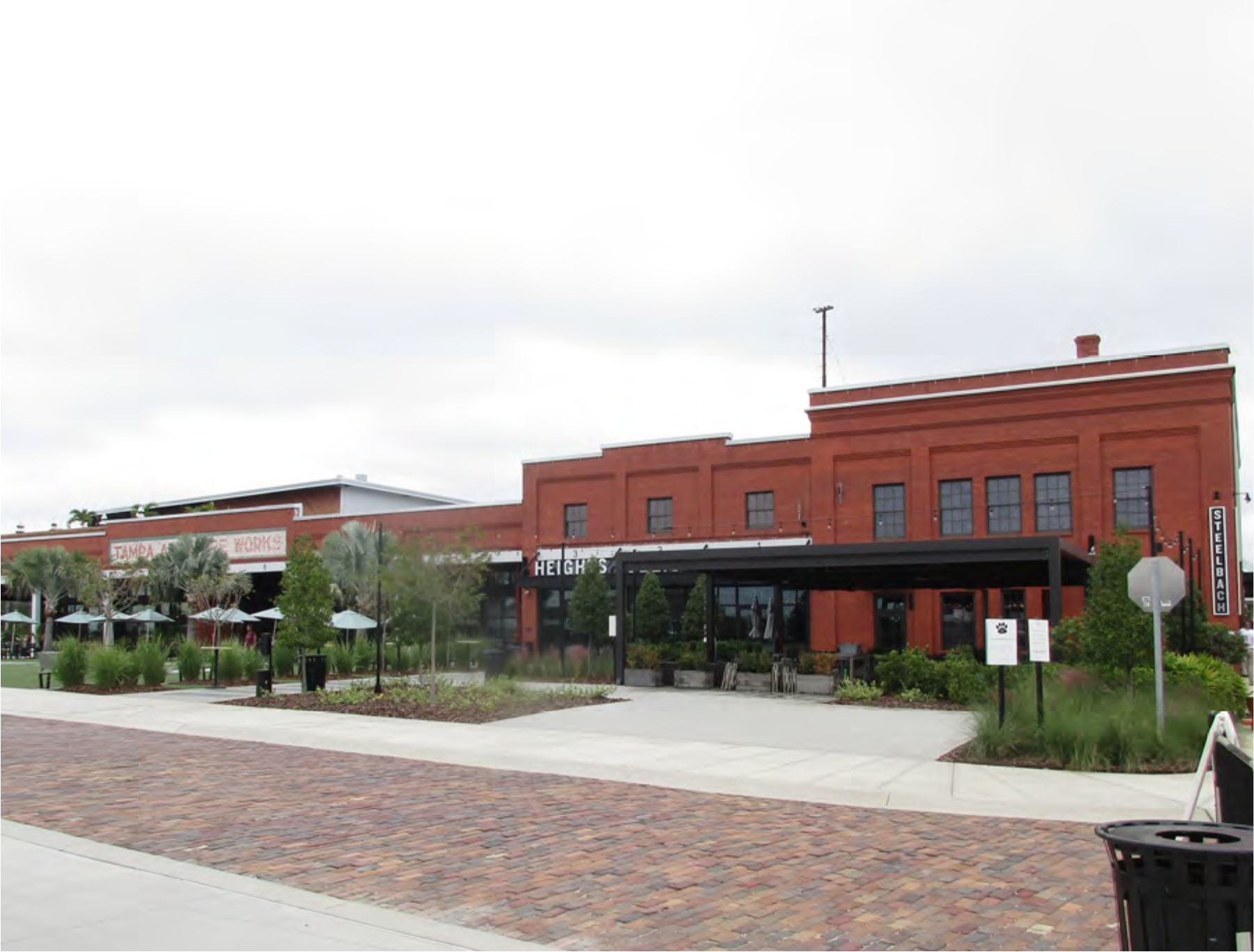 Exterior view of a brick restaurant building, with outdoor seating and nearby shops, under a cloudy sky.