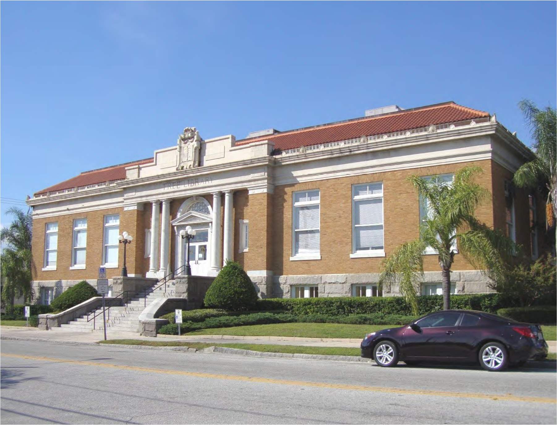 View of a historic brick building with columns and a red tile roof, surrounded by greenery and a parked car.