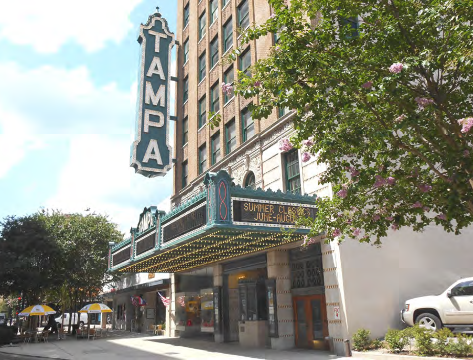 Historic Tampa Theatre with marquee, showcasing summer events, surrounded by trees and buildings under a blue sky.