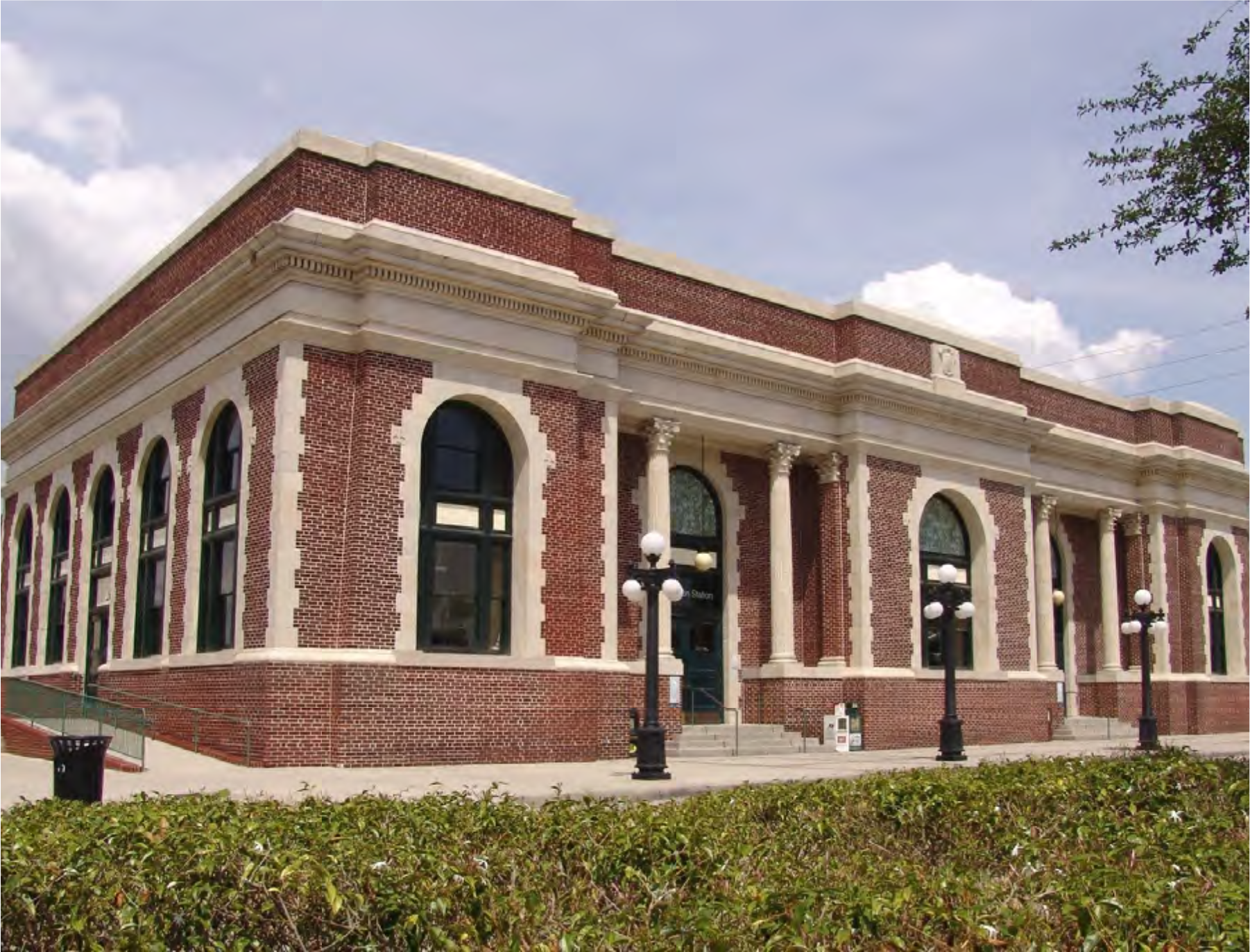 Historic brick building with large arched windows and decorative columns, surrounded by greenery and street lamps under a cloudy sky.
