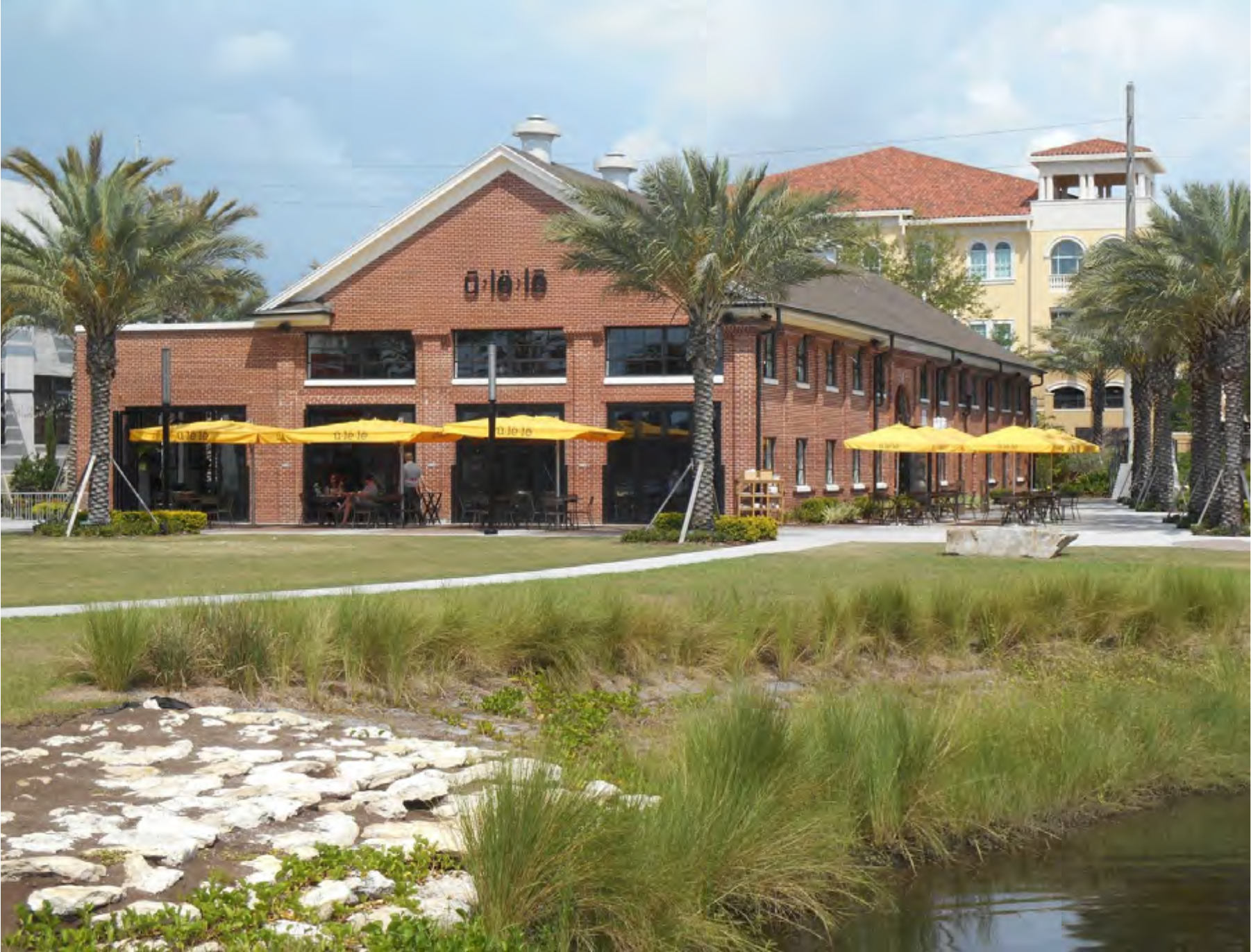 A brick building with outdoor seating and umbrellas, surrounded by palm trees and a small body of water.